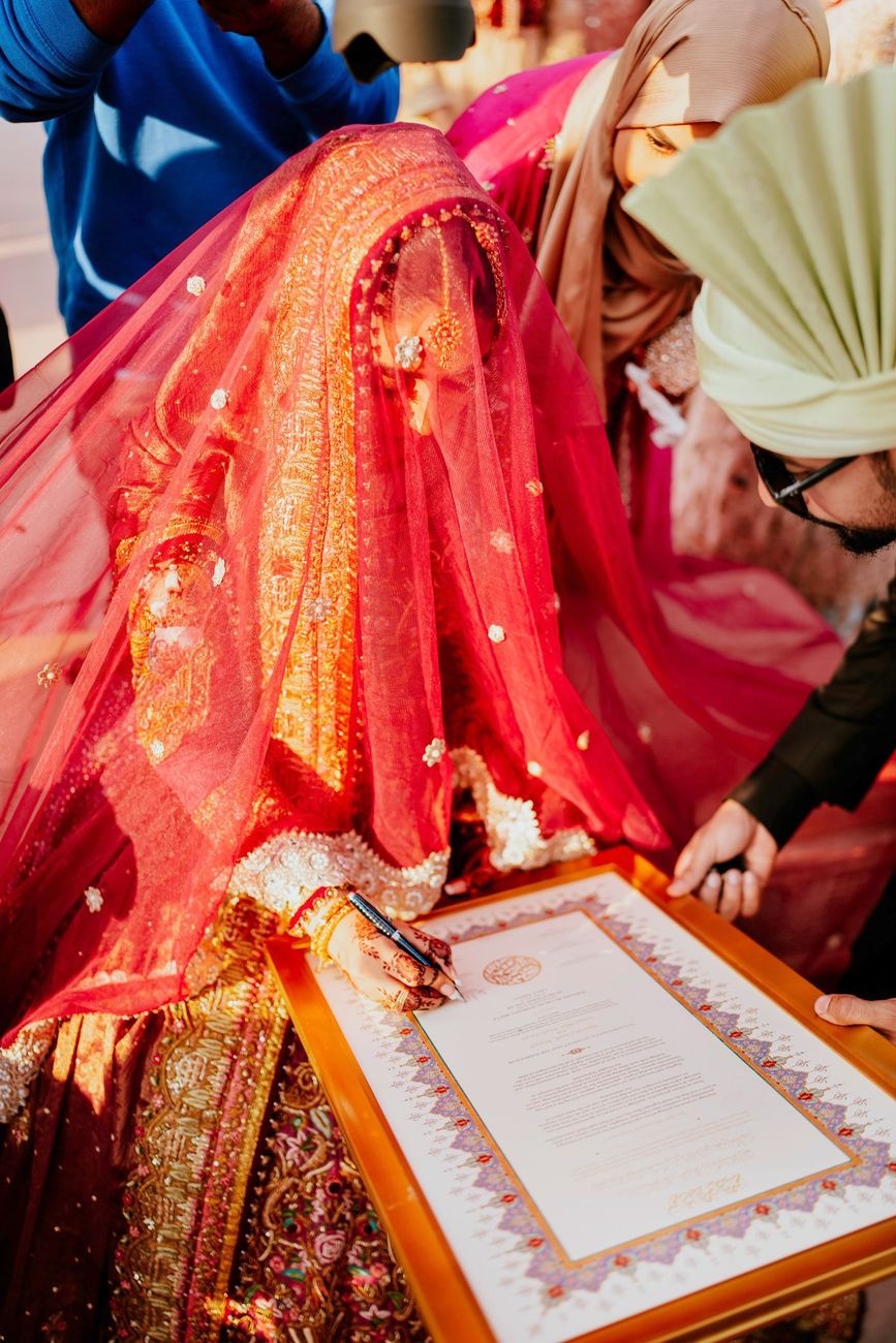 Bride signing the decorative nikah certificate, her face veiled, a family member leaning in to assist