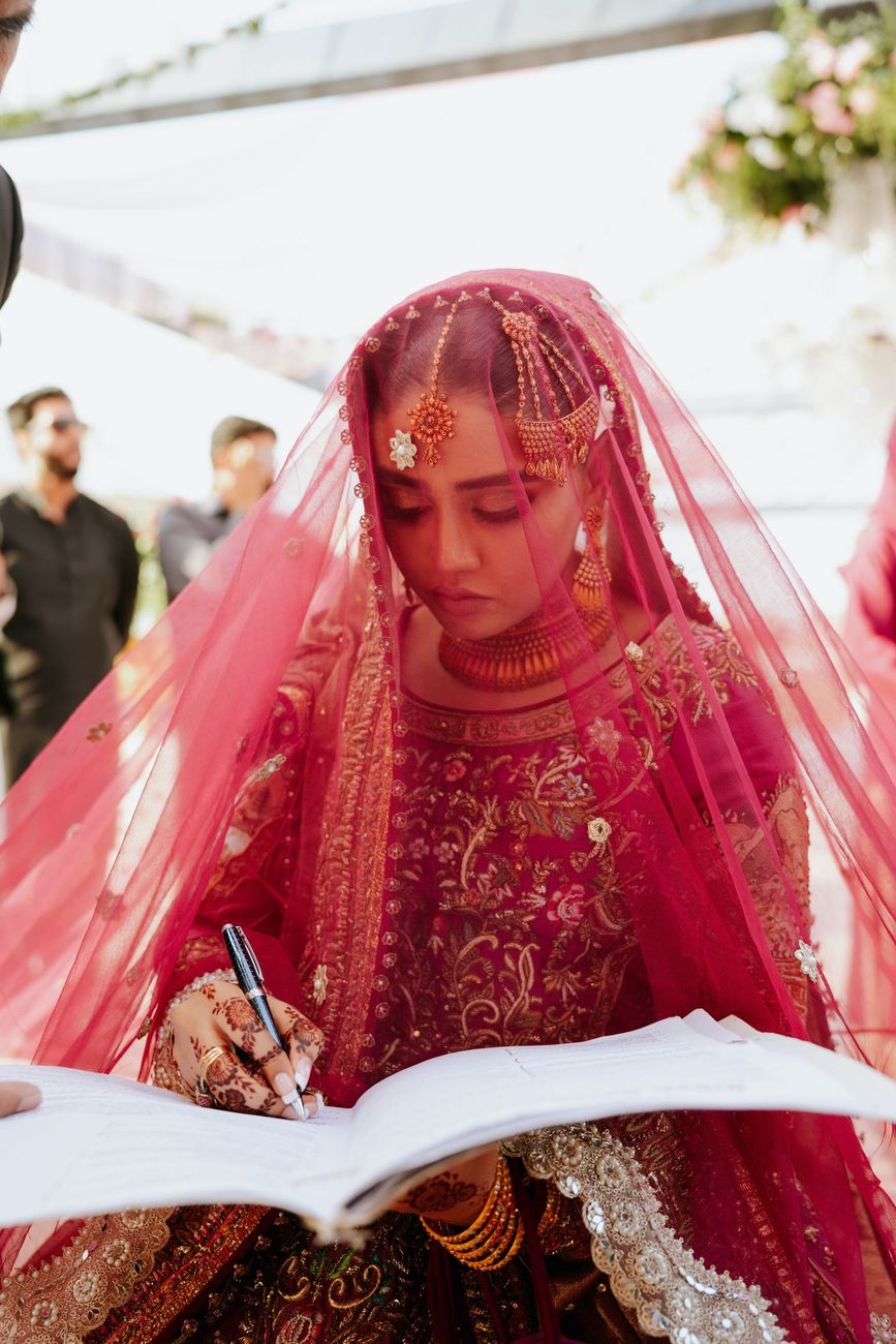 Close-up of the bride signing the nikah register, her face veiled beneath the embroidered red dupatta