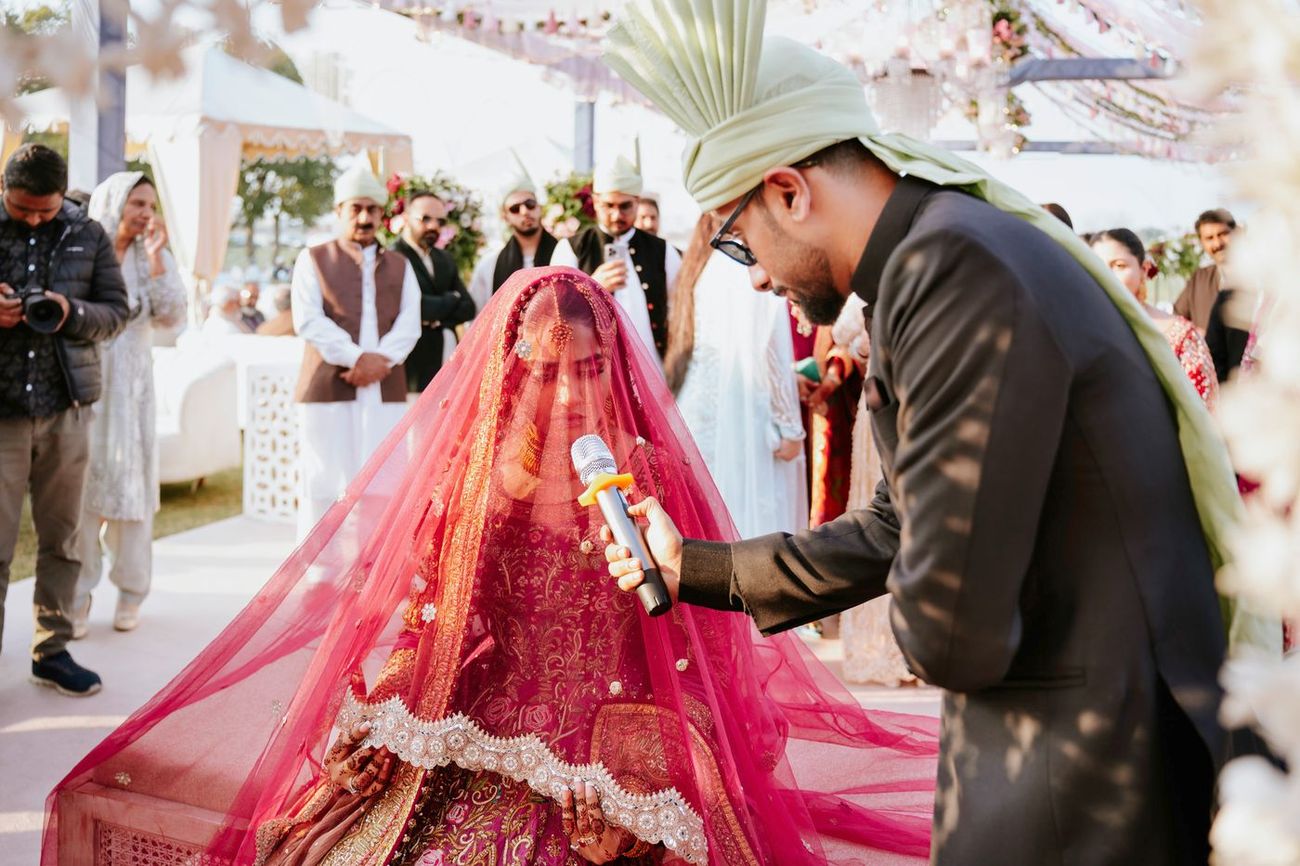 A guest holding a microphone to the veiled, seated bride as she answers during the nikah, photographer and turbaned guests looking on