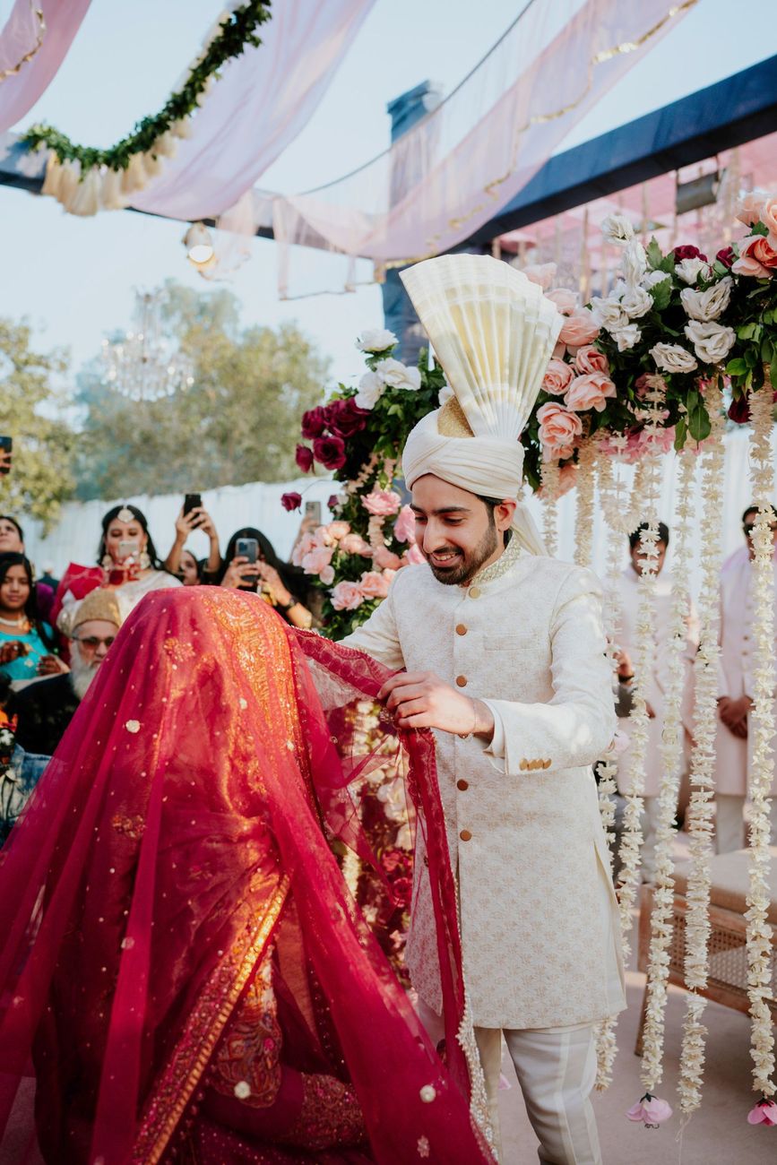Groom smiling as he lifts the bride's red veil after the nikah, floral garlands and a chandelier hanging behind them