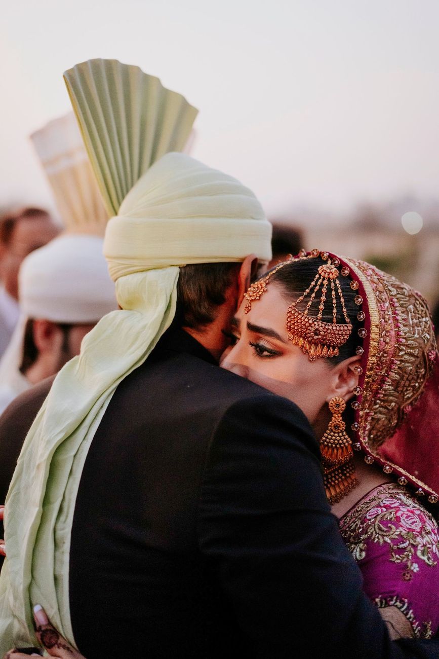Bride embracing a male relative in a black coat and pale green turban at the wedding venue