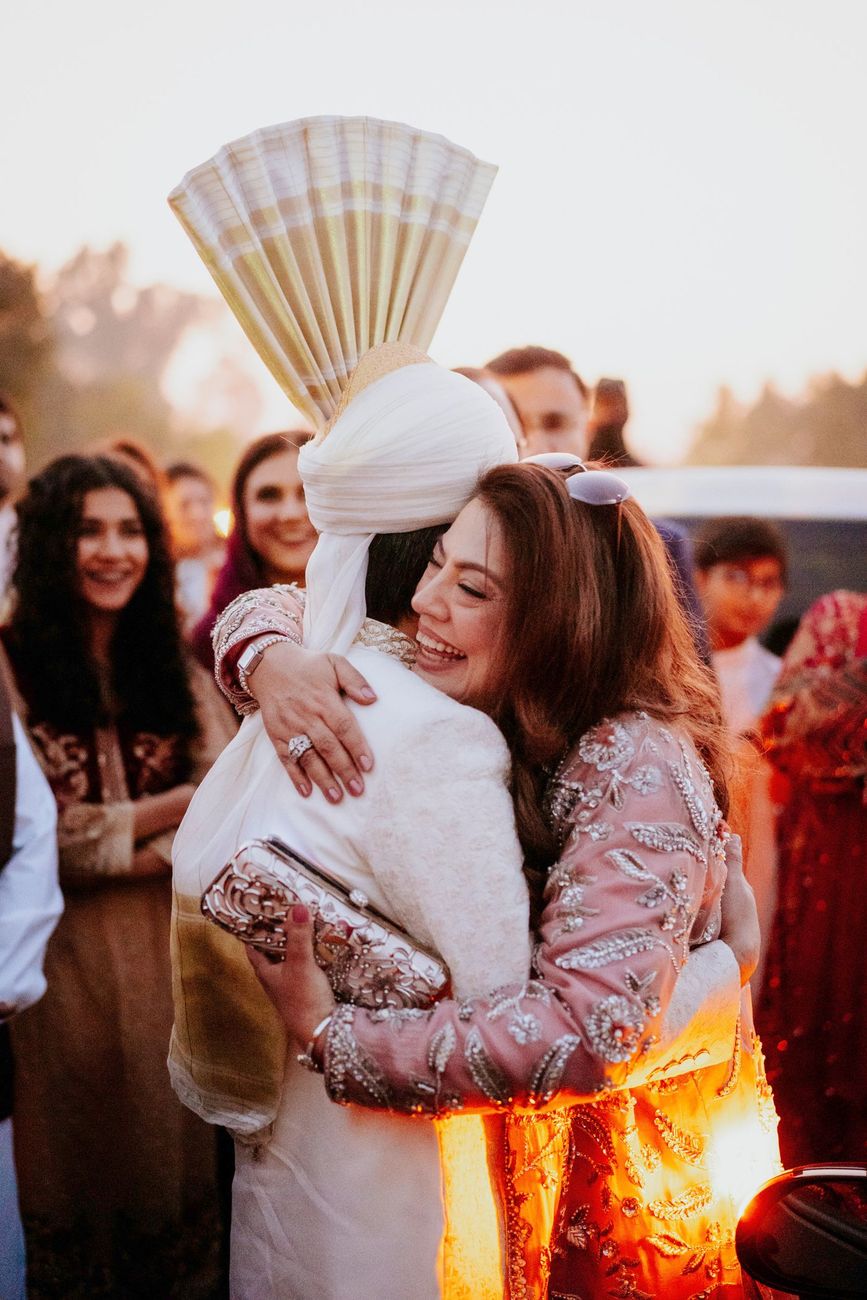 Groom in his ivory sherwani embracing a smiling female guest in a pink embellished outfit at golden hour