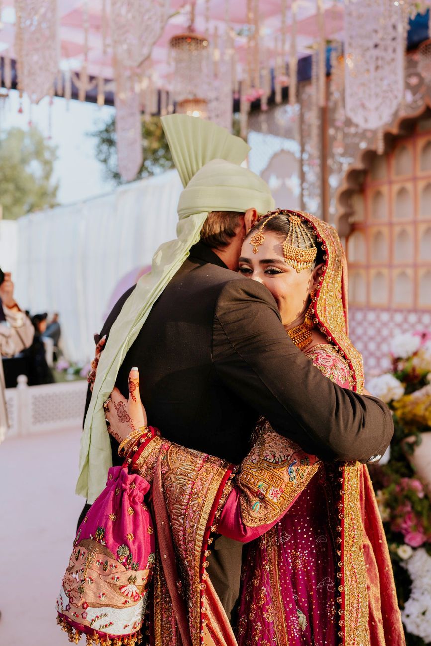 Bride embracing a male relative in a black coat and pale green turban beneath the venue's hanging crystal decor