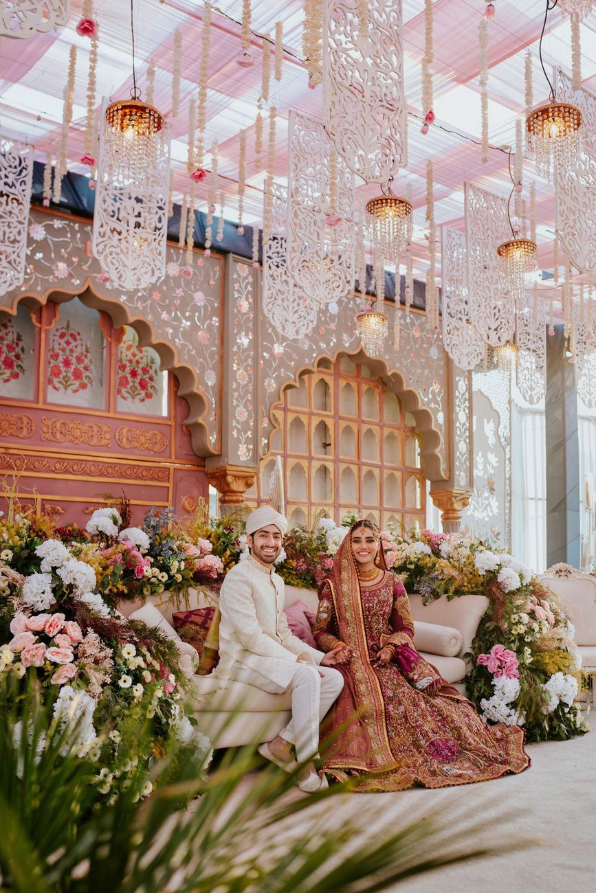 Bride and groom seated together on a sofa flanked by florals on the reception stage, its arched pink-and-gold Mughal-style backdrop and hanging lattice pendant lights behind them