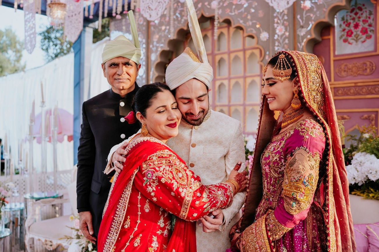 Bride and groom embracing the groom's parents on the reception stage, his father in a black coat and pankha turban, his mother in a red embroidered outfit