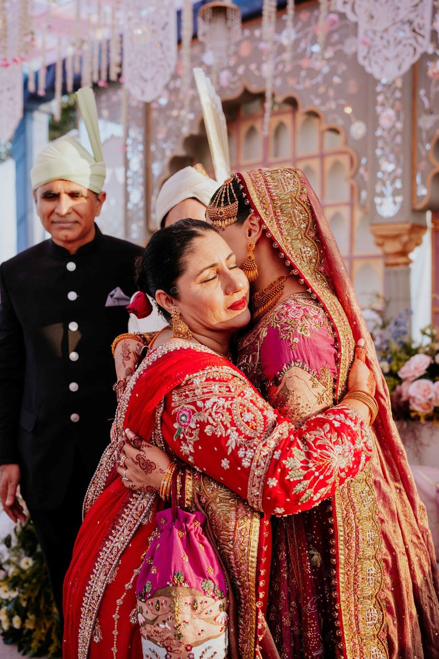 Bride embracing the groom's mother in a red embroidered outfit, his father standing behind them on the reception stage