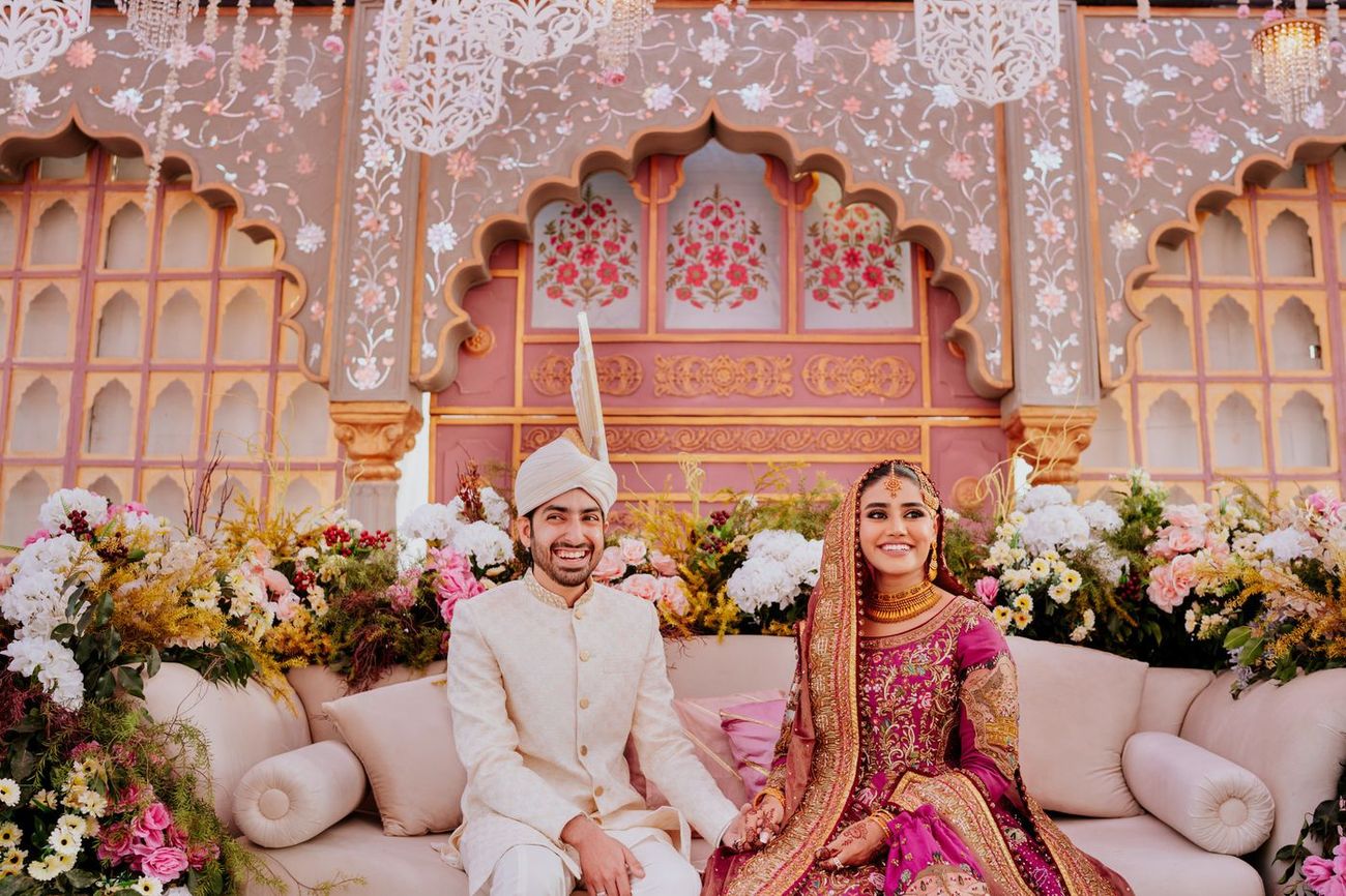 Bride and groom seated together on a floral-flanked sofa, smiling at the camera, reception stage backdrop behind them