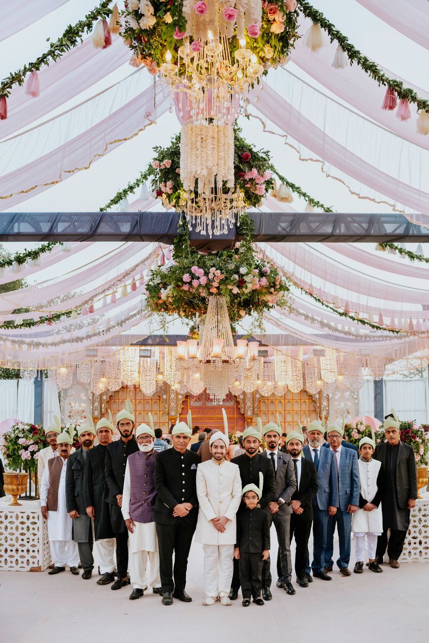 Wide shot of the groom with a large group of male relatives standing under the draped pink tent and hanging floral chandeliers, reception stage visible behind them