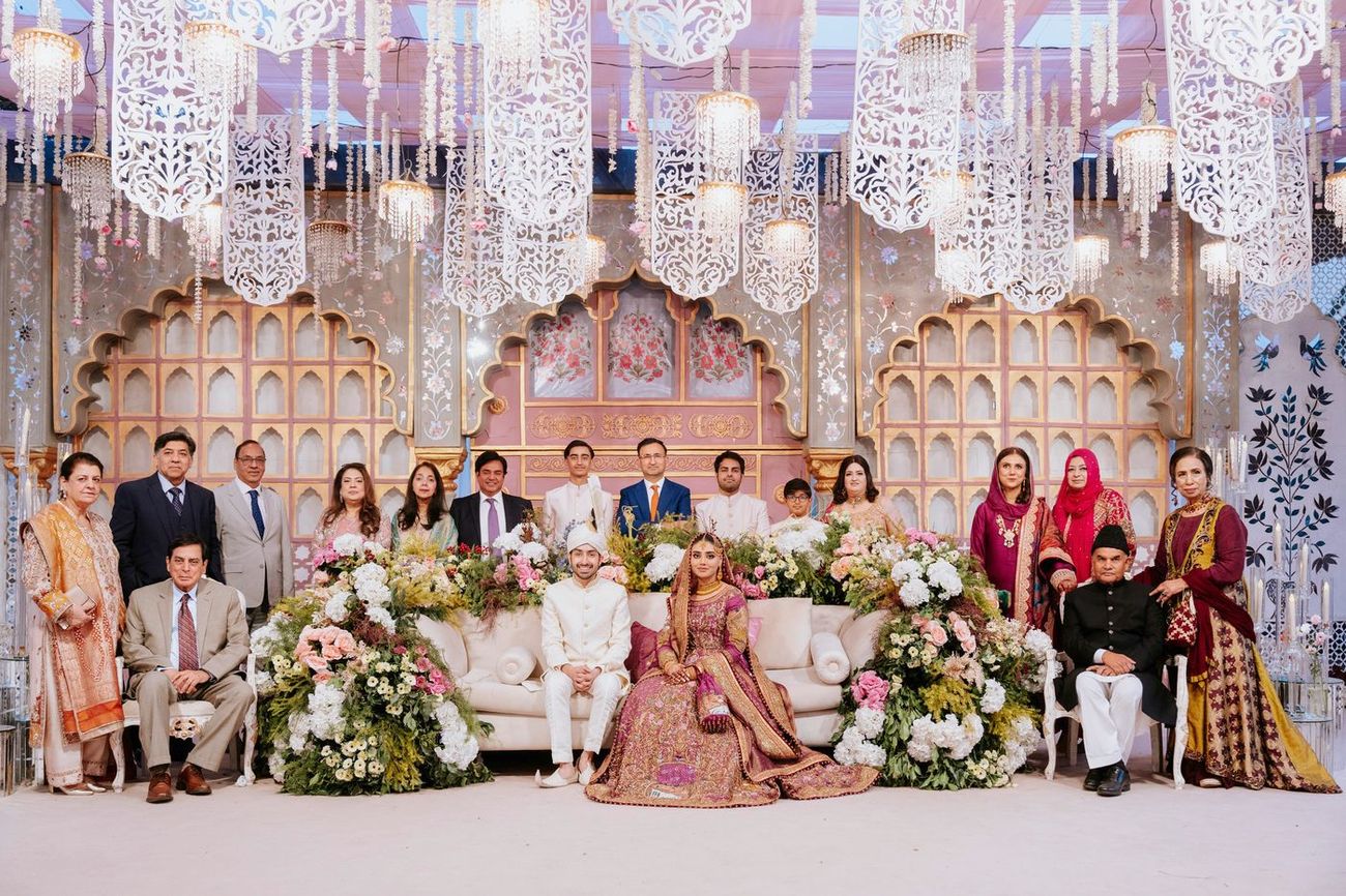 Extended family group portrait with the bride and groom seated centrally on a floral-decorated sofa, full reception stage backdrop behind them