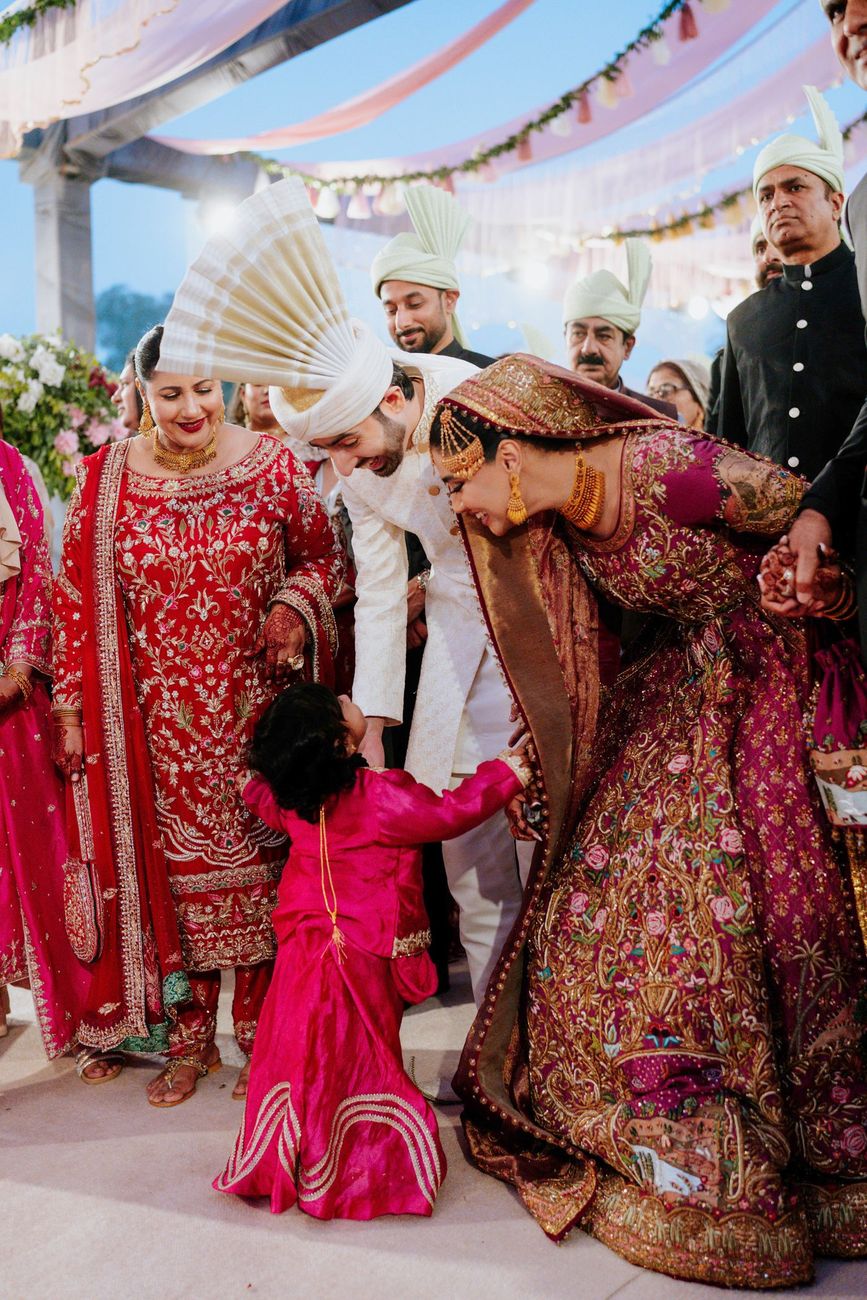 Bride and groom bending down to greet a young girl in a pink outfit, family members looking on