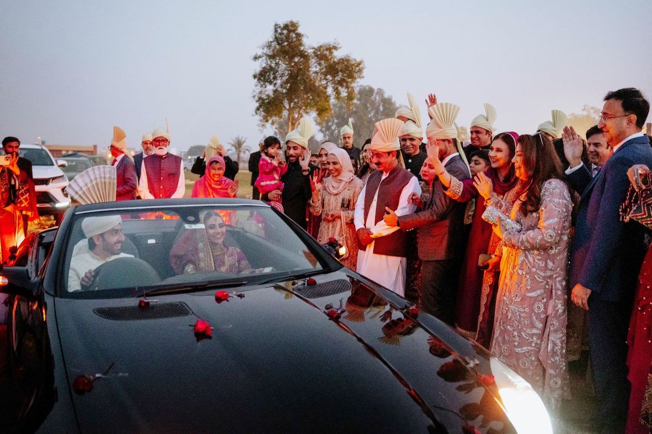 Wide shot of the bride and groom's send-off in a rose-petal-strewn convertible, family and guests waving on either side at dusk