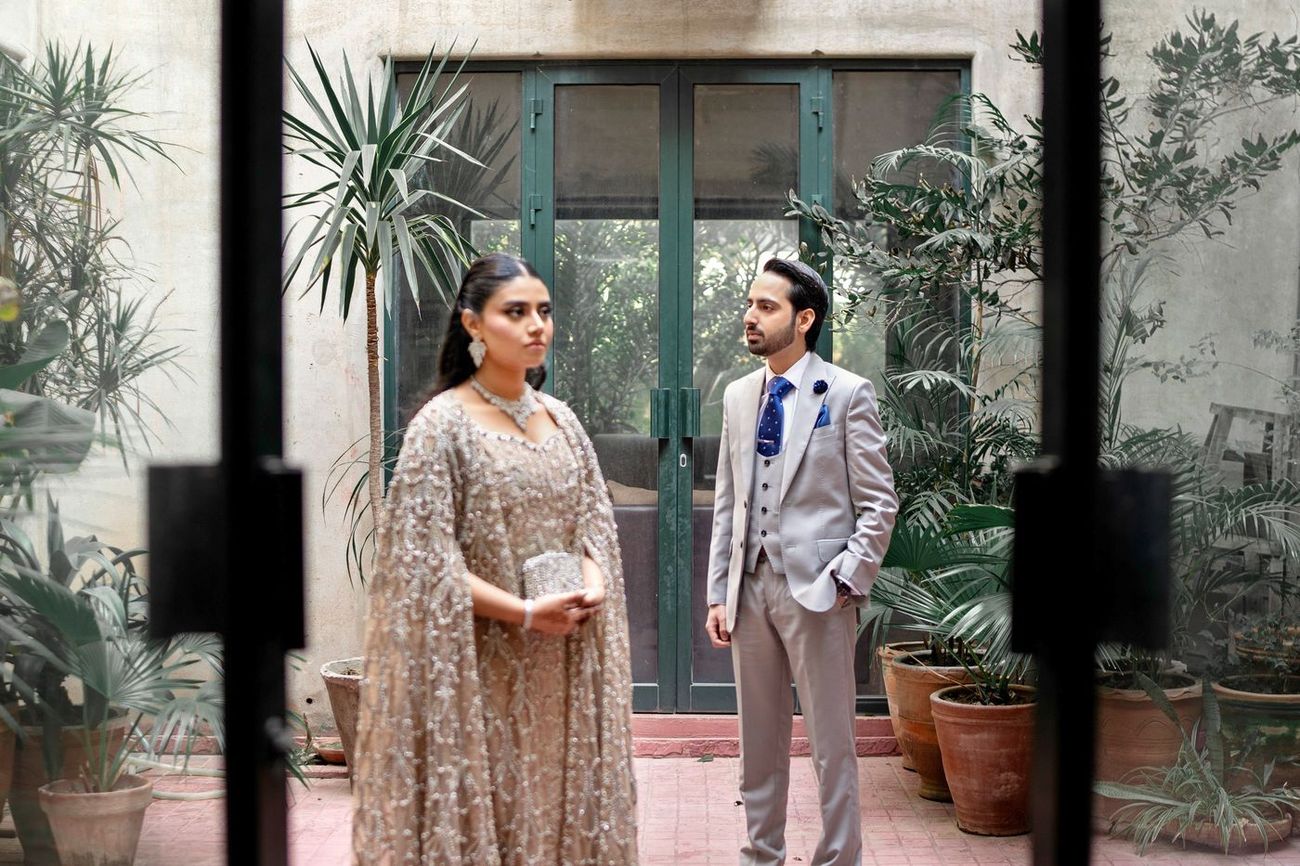 Couple glimpsed through a doorway, the bride in a beaded champagne gown and the groom in a grey suit, framed by potted plants