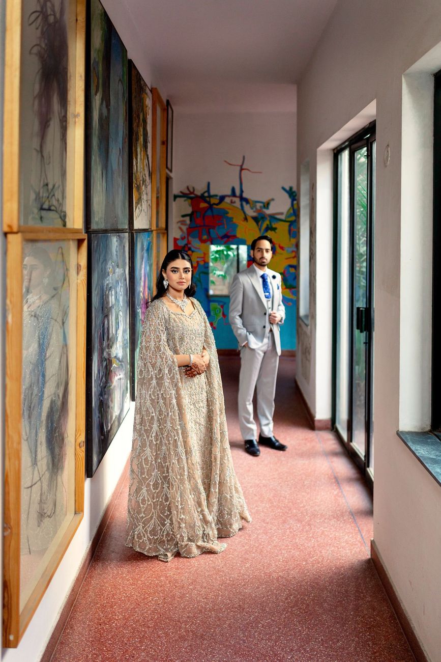 Bride standing in an art-studio corridor lined with paintings, groom standing behind her