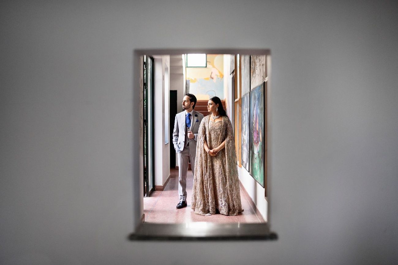 Couple framed through a small rectangular opening in a wall, standing together in the art-studio hallway