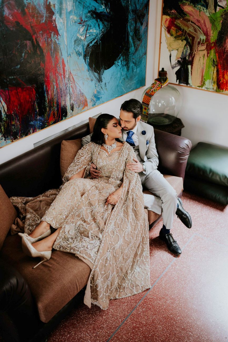 Wide shot of the couple seated together on a sofa beneath bold abstract artwork, a patterned ceramic jar beside them