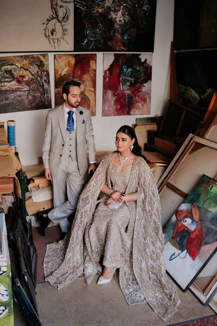 Groom standing beside the seated bride in the cluttered art studio, surrounded by paintings and easels
