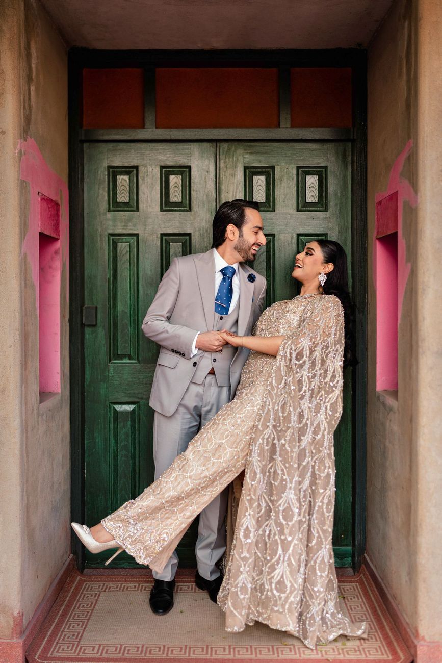 Bride and groom laughing together in a doorway framed by a green wooden door, she playfully lifting one leg