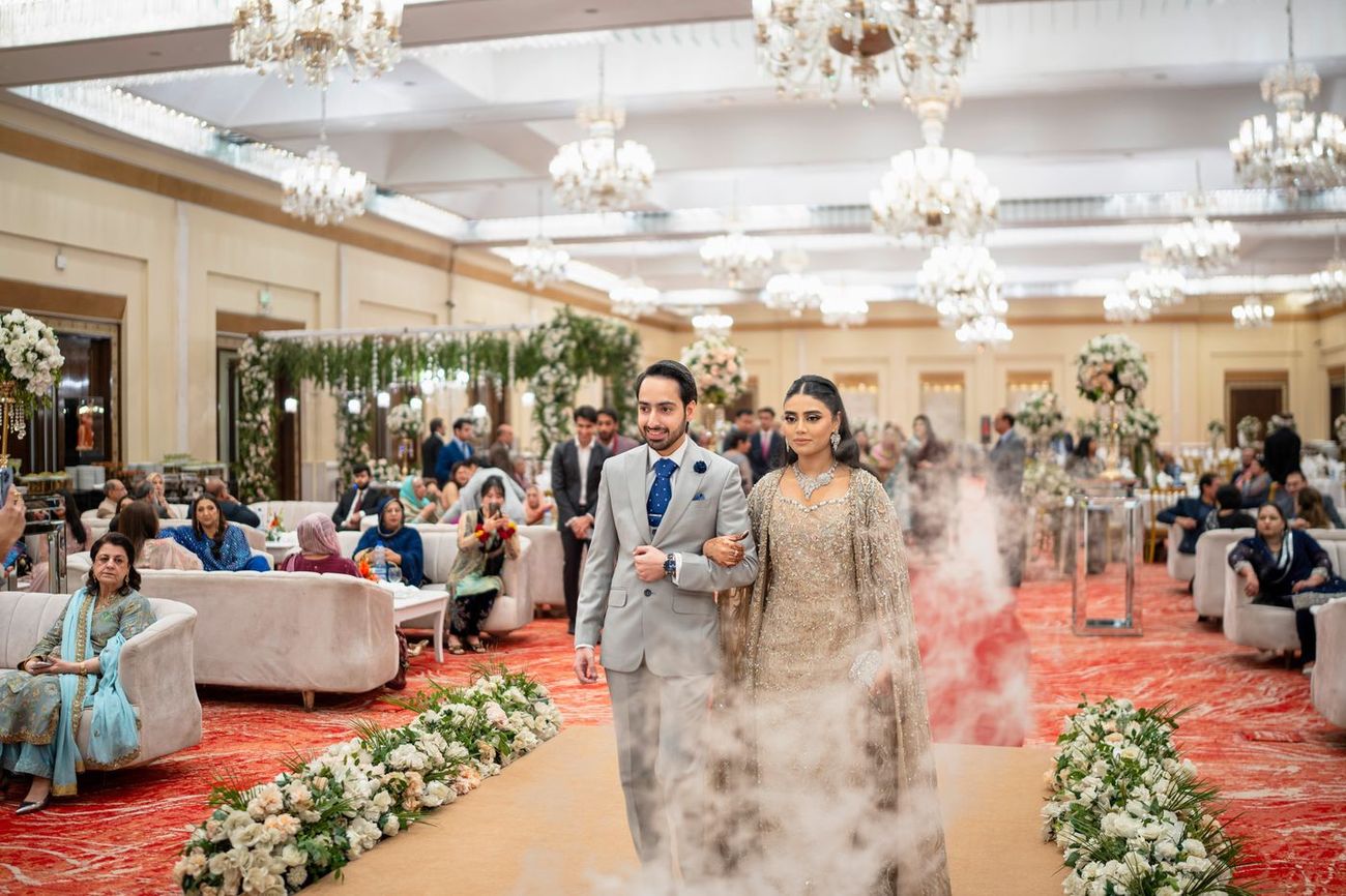 Wide shot of the bride and groom walking through a fog-covered aisle in the ballroom, guests seated on either side beneath crystal chandeliers