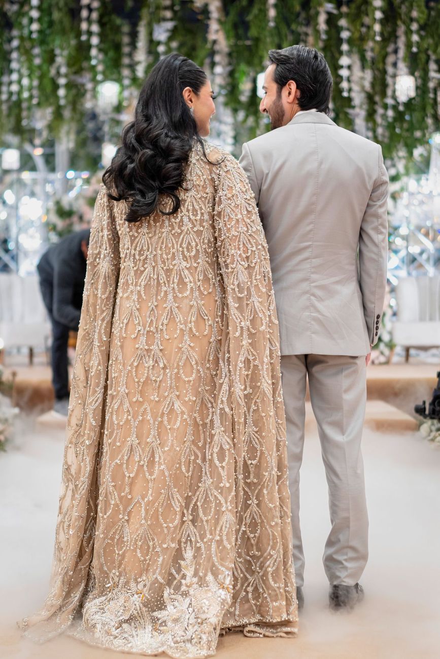Bride and groom seen from behind, walking toward a hanging floral-and-crystal installation on the reception stage