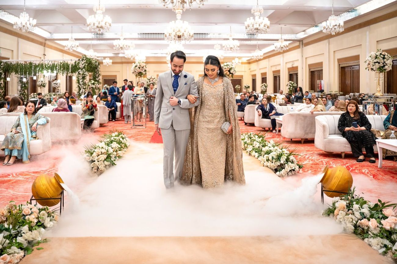 Wide shot of the bride and groom walking through smoke-filled urns lining the ballroom aisle, guests seated on either side
