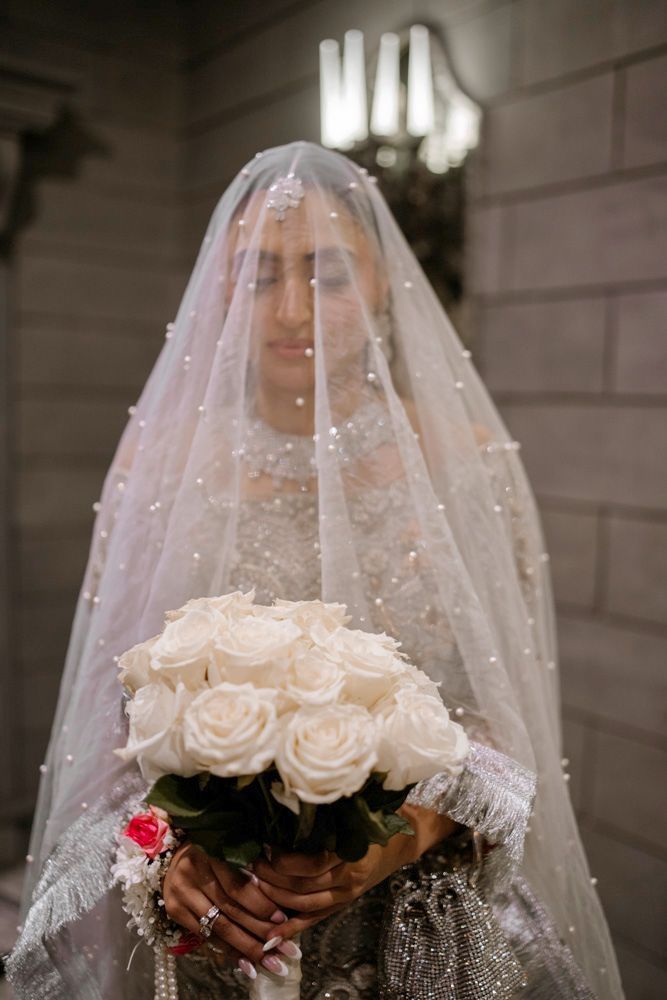 Bride beneath her pearl-beaded veil holding a bouquet of white roses, Sheraz & Nawal's wedding at Nishat Hotel, Lahore