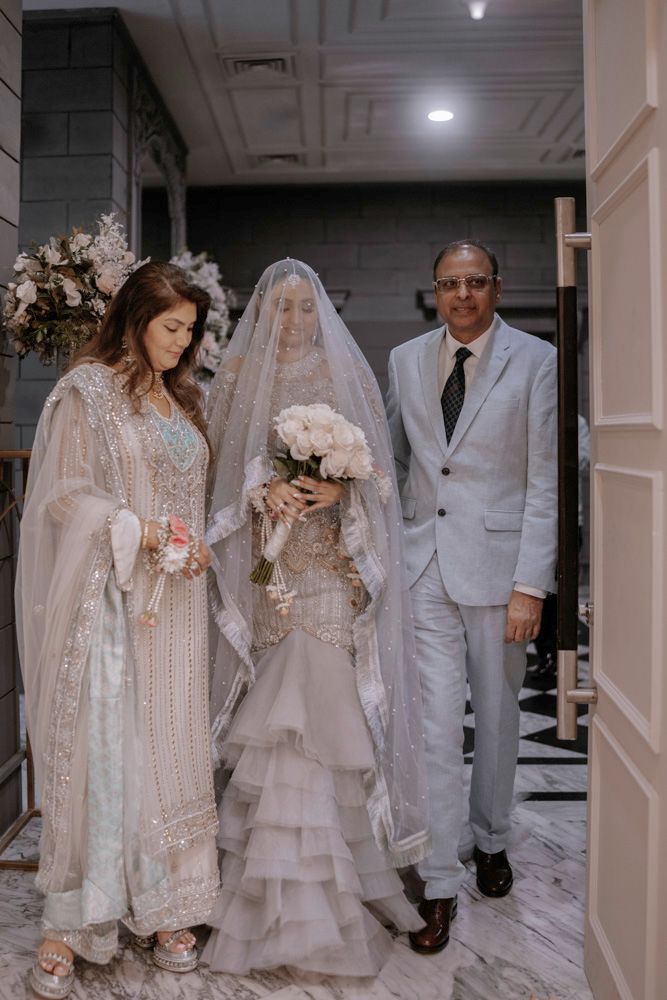 Bride's entrance with her parents through a marble hallway, holding a white rose bouquet, Sheraz & Nawal's wedding at Nishat Hotel, Lahore