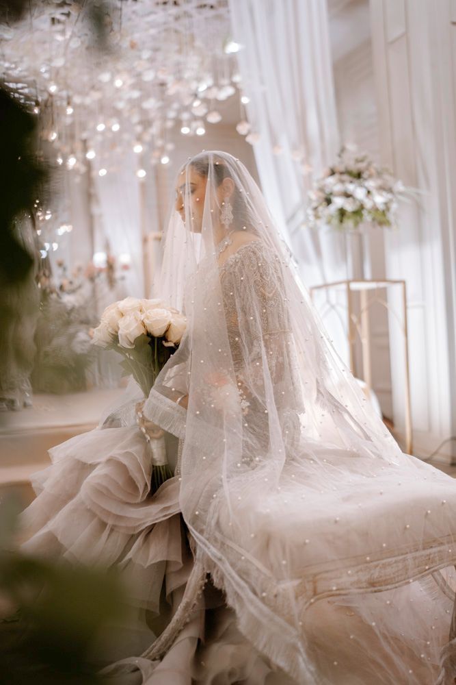 Bride seated on a tufted bench beneath her veil, holding a rose bouquet beside floral decor, Sheraz & Nawal's wedding at Nishat Hotel, Lahore