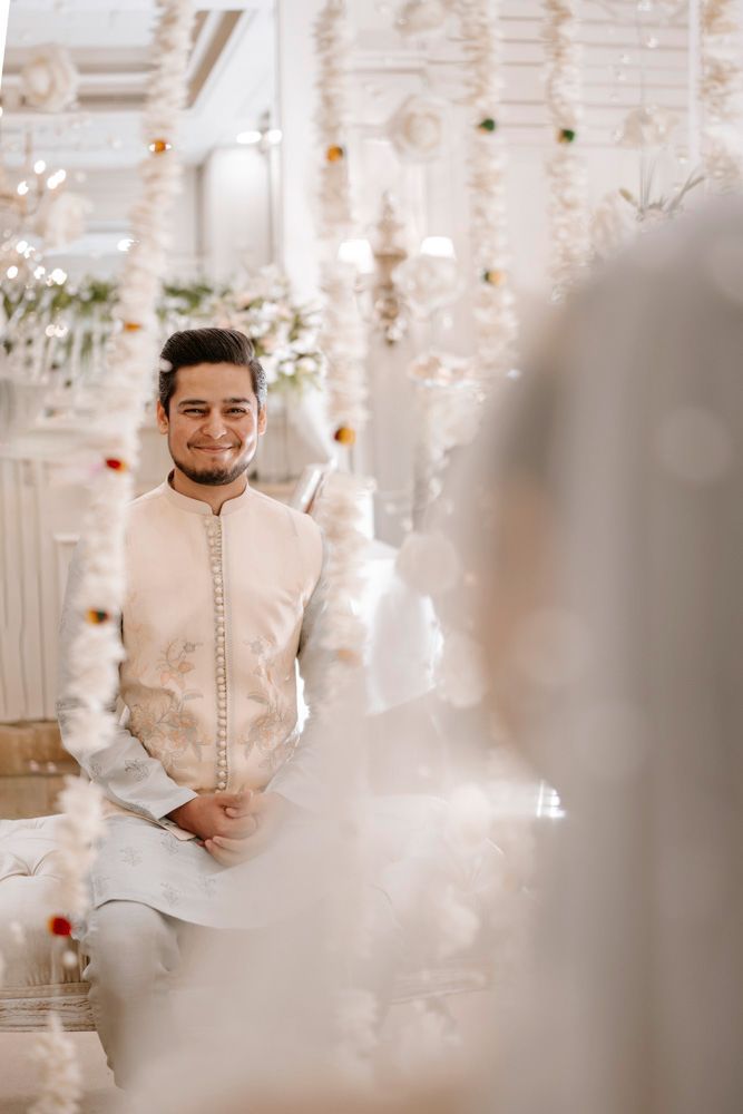 Groom smiling, seated on a tufted bench, viewed through a hanging floral curtain, Sheraz & Nawal's wedding at Nishat Hotel, Lahore