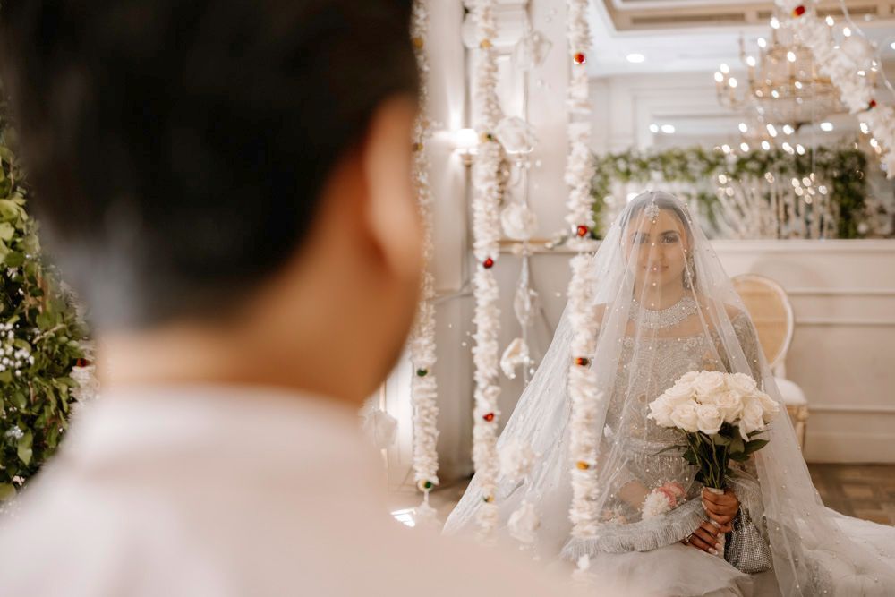 Bride beneath her veil seated on a chair, viewed past the groom's shoulder, Sheraz & Nawal's wedding at Nishat Hotel, Lahore