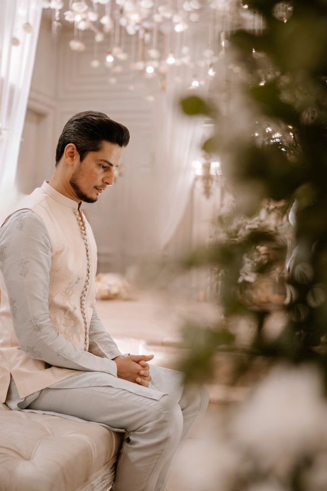 Solo groom portrait seated in profile, hands clasped, soft chandelier light behind him, Sheraz & Nawal's wedding at Nishat Hotel, Lahore