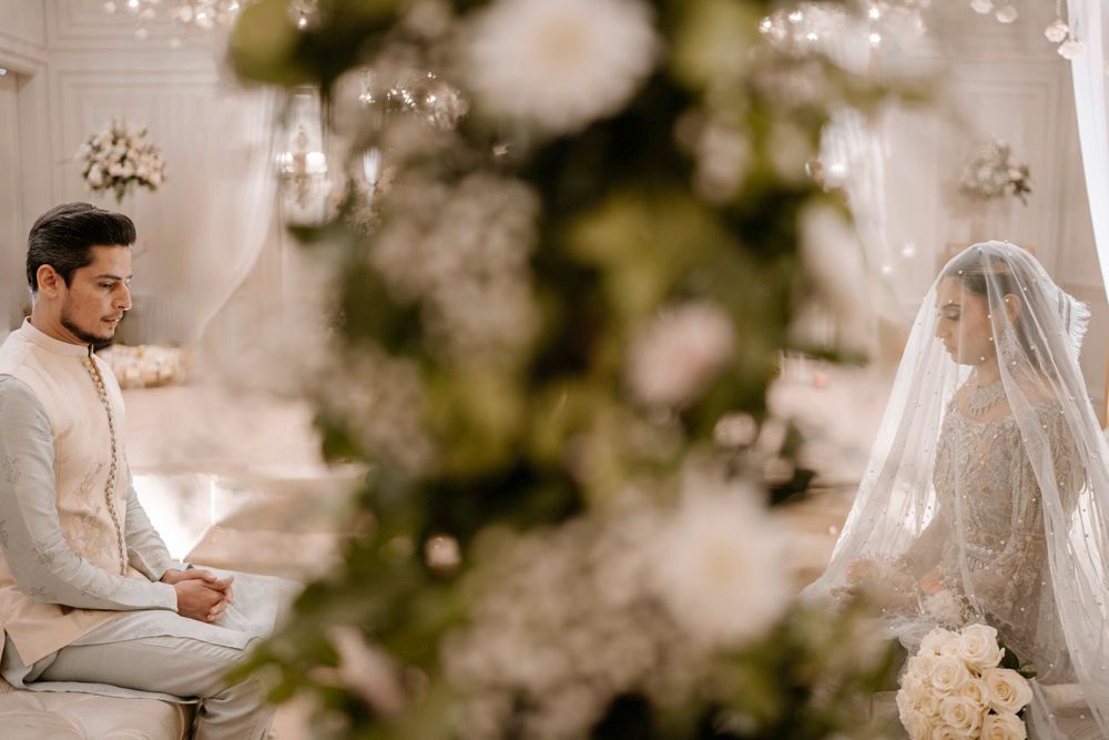 Bride and groom seated for the nikah, viewed through a blurred floral foreground, Sheraz & Nawal's wedding at Nishat Hotel, Lahore