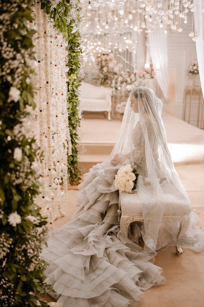 Bride seated beneath her veil on a tufted bench, viewed through hanging floral garlands, Sheraz & Nawal's wedding at Nishat Hotel, Lahore