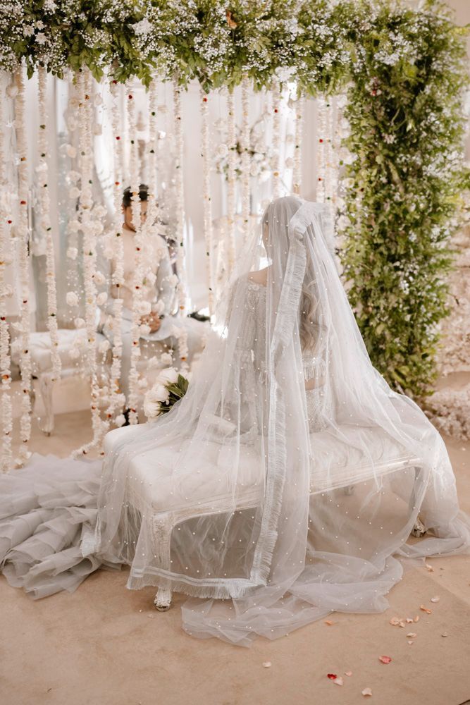 Bride viewed from behind seated on a tufted bench, groom visible through the floral curtain behind her, Sheraz & Nawal's wedding at Nishat Hotel, Lahore