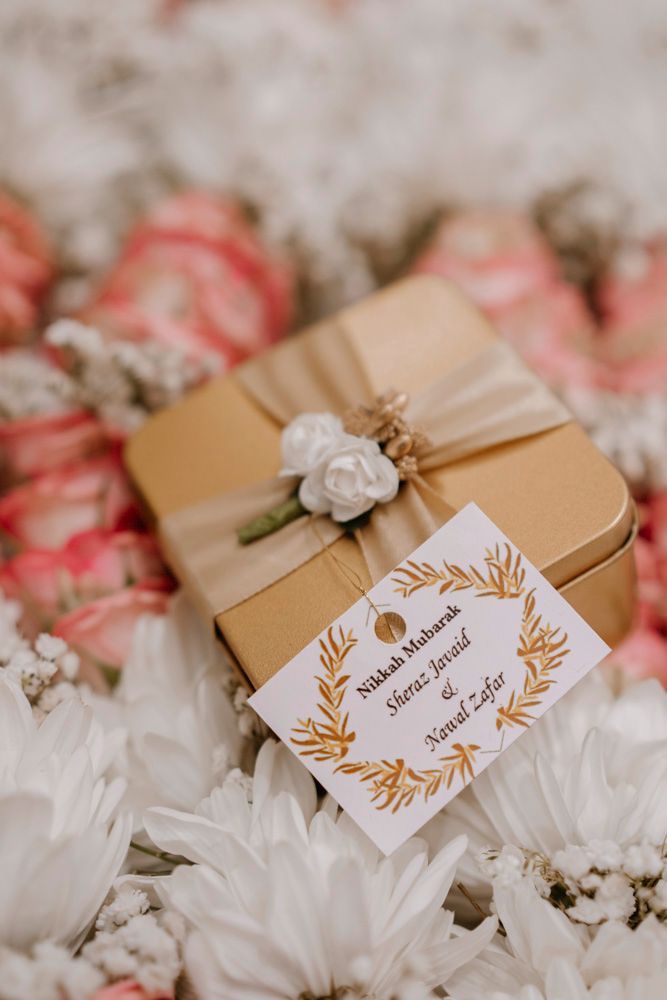 Detail of a gold nikah favor box tied with ribbon and a wedding tag reading Nikkah Mubarak, Sheraz & Nawal's wedding at Nishat Hotel, Lahore