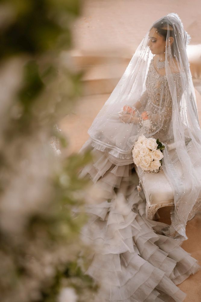 Bride seated in profile on a tufted bench, holding a white rose bouquet, ruffled train pooling below, Sheraz & Nawal's wedding at Nishat Hotel, Lahore