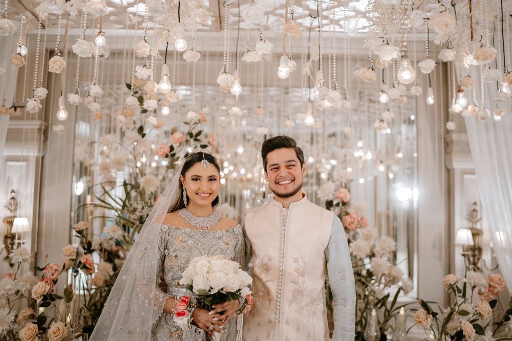 Close-up portrait of bride and groom smiling together beneath hanging floral lights, Sheraz & Nawal's wedding at Nishat Hotel, Lahore