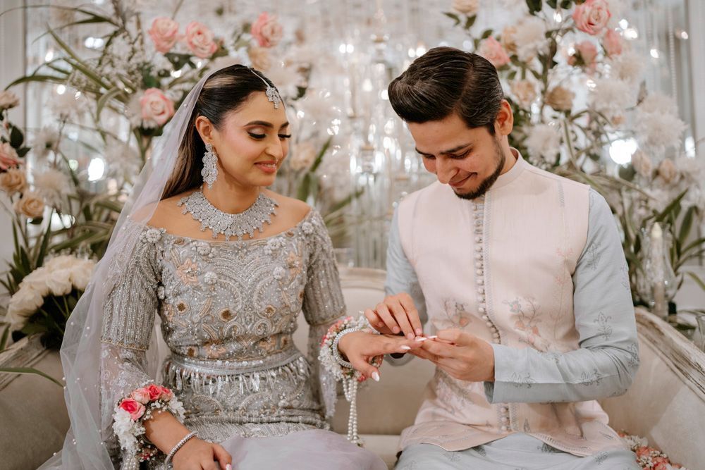 Groom placing a ring on the bride's finger as both smile, floral chandeliers behind them, Sheraz & Nawal's wedding at Nishat Hotel, Lahore