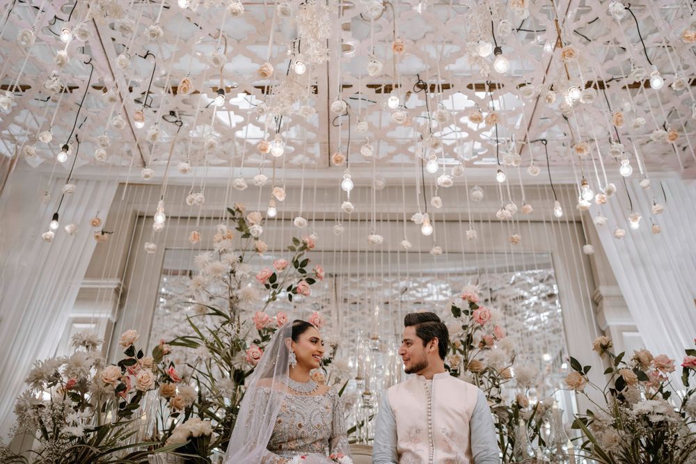 Bride and groom seated together smiling, framed by a lattice ceiling hung with roses and lights, Sheraz & Nawal's wedding at Nishat Hotel, Lahore