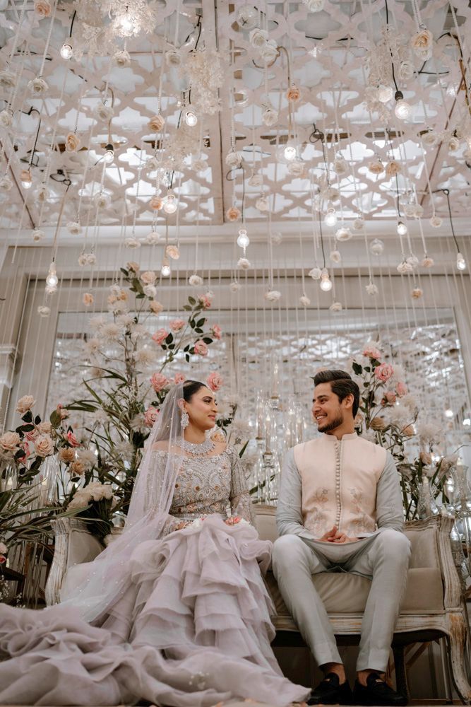Bride and groom seated together on a sofa beneath floral chandeliers, smiling at each other, Sheraz & Nawal's wedding at Nishat Hotel, Lahore