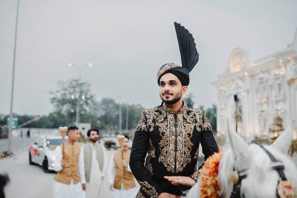 Groom on horseback during the baraat approaching the illuminated venue facade, Zara & Shuaib's wedding at Garrison Halls, Lahore