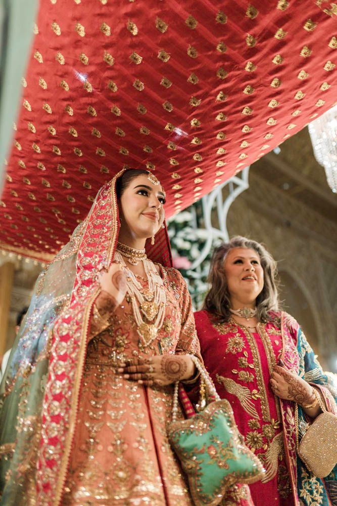 Bride's entrance beneath a red ceremonial cloth canopy, her mother walking beside her, Zara & Shuaib's wedding, Lahore