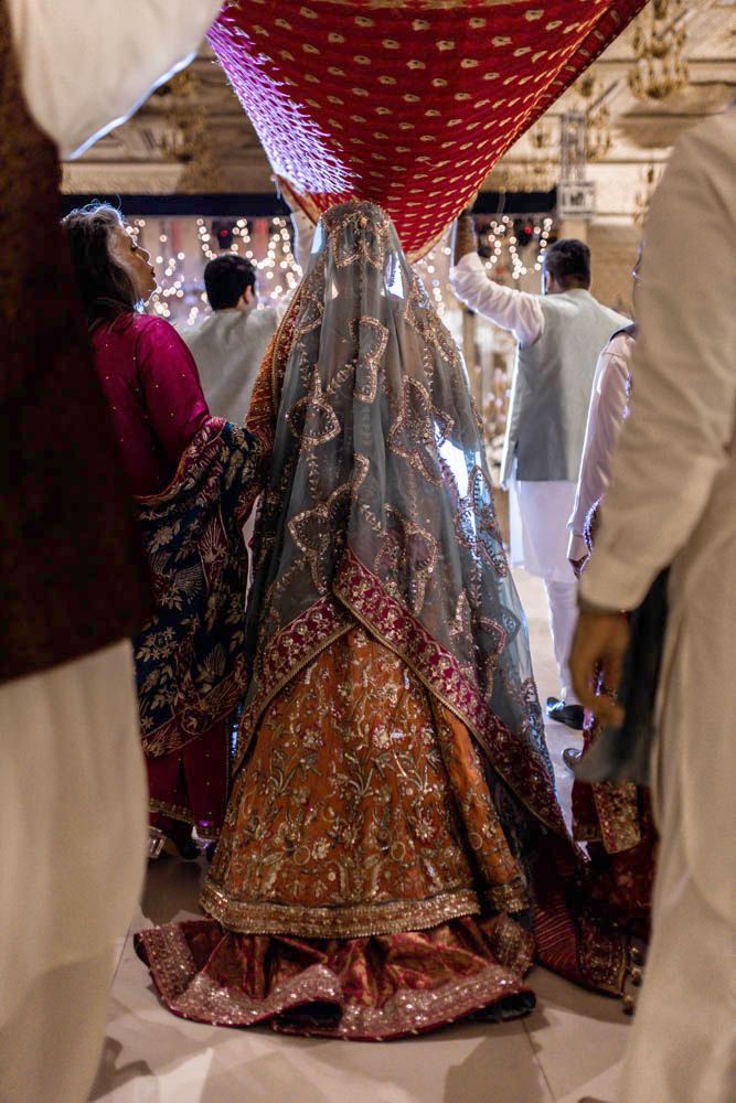 Bride viewed from behind walking under the red entrance canopy, her embellished orange lehenga visible, Zara & Shuaib's wedding, Lahore