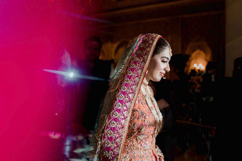 Close-up profile portrait of the bride in a pink and orange dupatta lit by colorful stage lighting, Zara & Shuaib's wedding, Lahore
