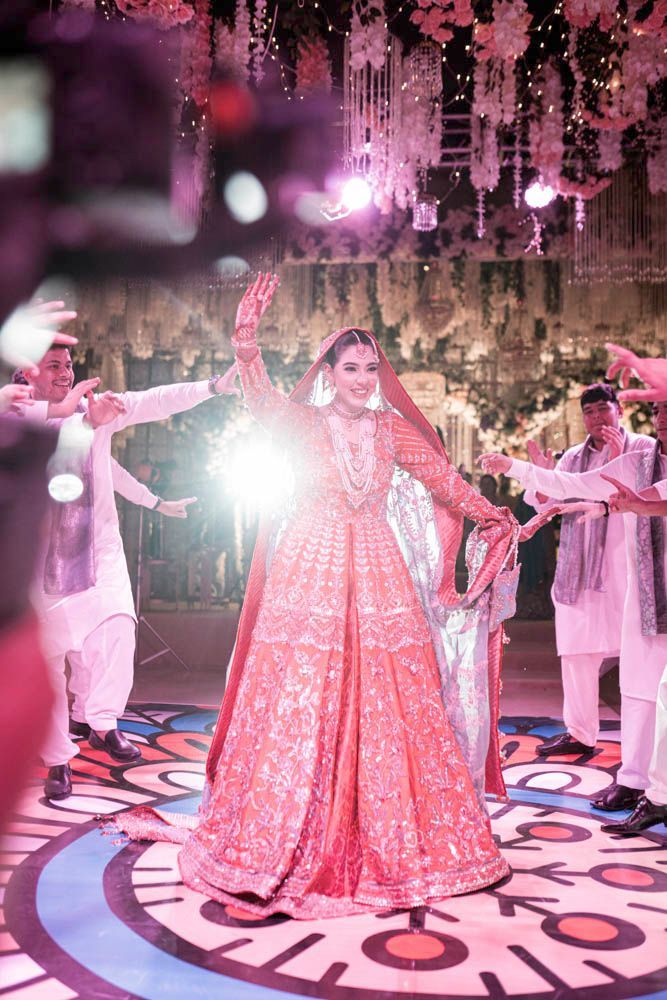 Bride dancing on the patterned reception dance floor in a coral gown, friends cheering around her, Zara & Shuaib's wedding at Garrison Halls, Lahore