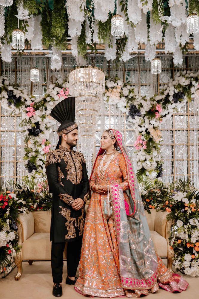 Bride and groom standing together in front of a chandelier and floral wall on the reception stage, Zara & Shuaib's wedding at Garrison Halls, Lahore