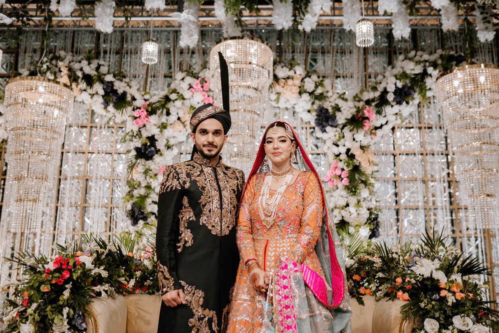 Bride and groom posing together beneath hanging crystal chandeliers and floral garlands, Zara & Shuaib's wedding at Garrison Halls, Lahore