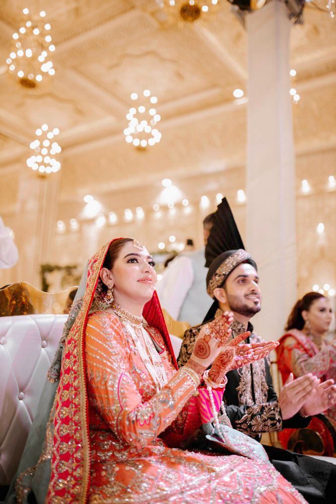 Bride and groom seated together clapping along to a performance, Zara & Shuaib's wedding at Garrison Halls, Lahore