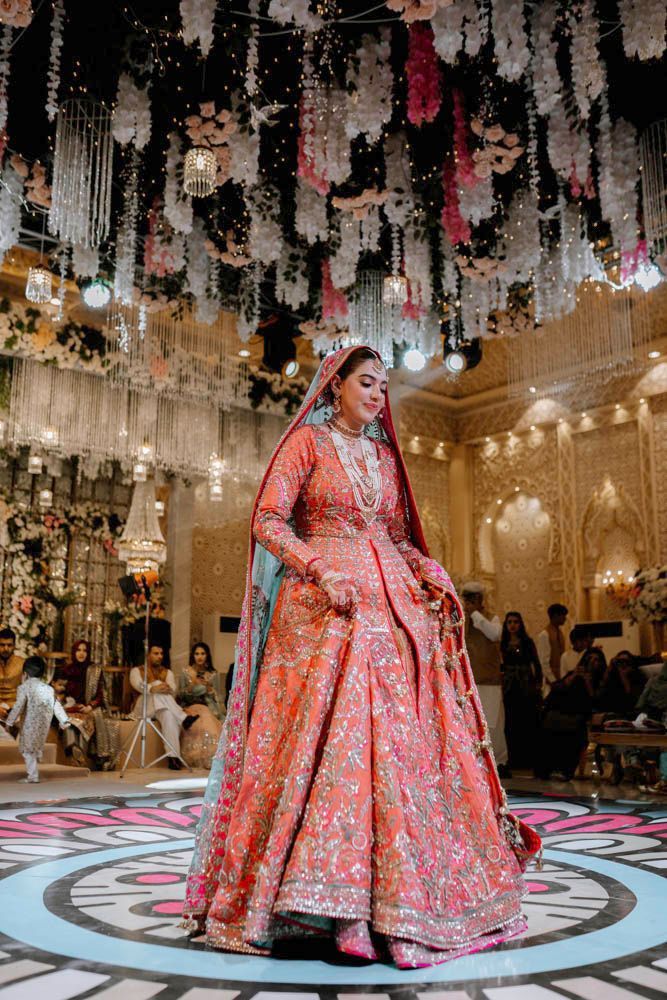 Solo bride dancing on the patterned floor in her coral lehenga, beneath floral chandeliers, Zara & Shuaib's wedding at Garrison Halls, Lahore