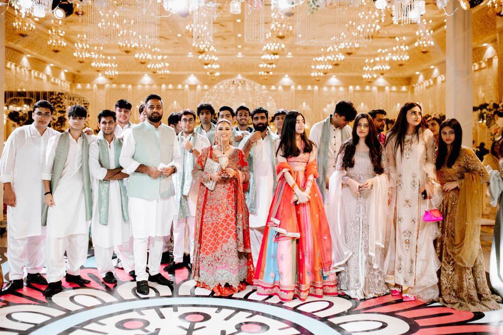Large group photo of bride's friends and family on the dance floor beneath grand chandeliers, Zara & Shuaib's wedding at Garrison Halls, Lahore