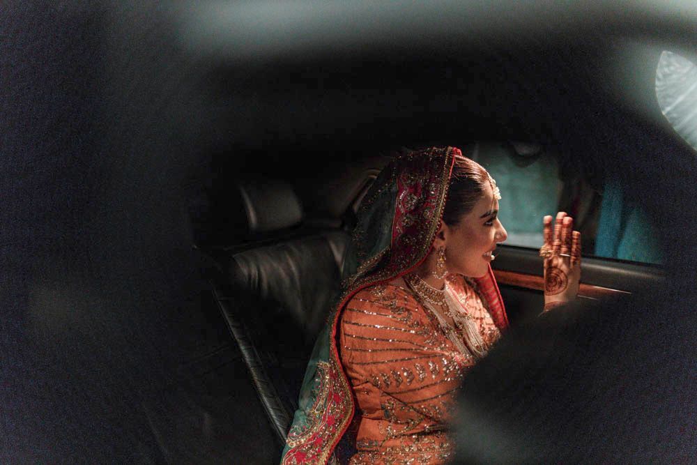 Bride waving from inside a departure car at night, seen through the car window, Zara & Shuaib's wedding, Lahore